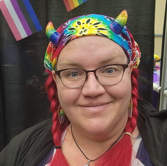 A photo of Kari, a woman wearing glasses, a tie-dye bandana, and rainbow horns. She has red braids on both sides, and a welcoming smile.